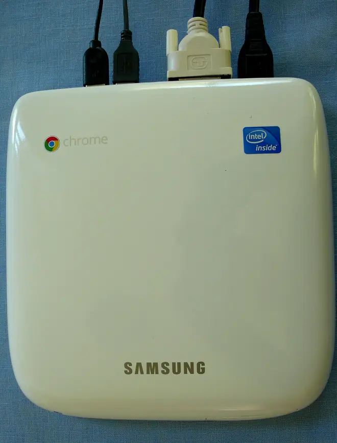 White compact Chromebox computer with the Chrome logo and display cables plugged in, standing upright on a desk
