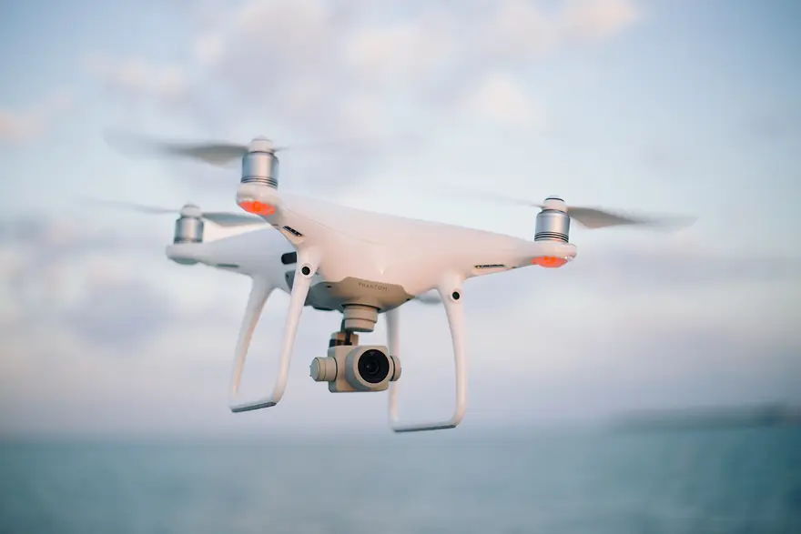White camera drone hovering in flight against soft clouds above open water