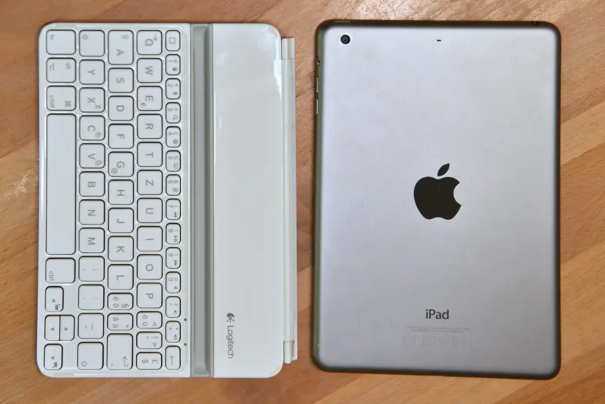 Silver Apple iPad mini lying face down beside a compact white keyboard on a wooden desk